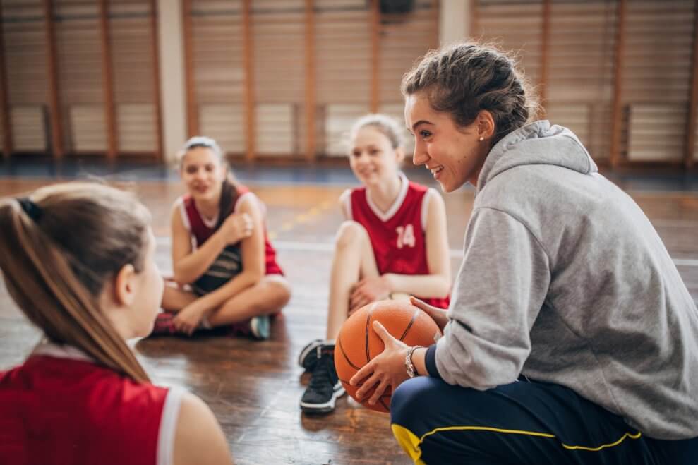 A coach sits on a basketball court with her female athletes sitting around her in a circle.