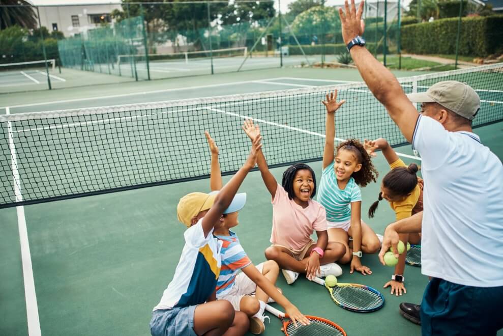 A tennis coach and his athletes sit on an outdoor court and do a cheer together.