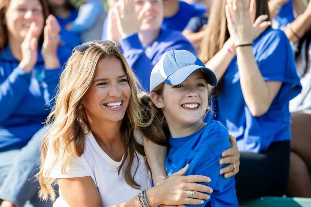 A mom and her daughter hug while cheering on the sidelines at a sports game.