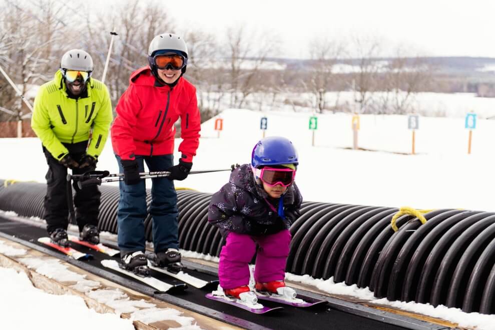 A dad, mom, and their young daughter ride a magic carpet ski lift at a ski resort.