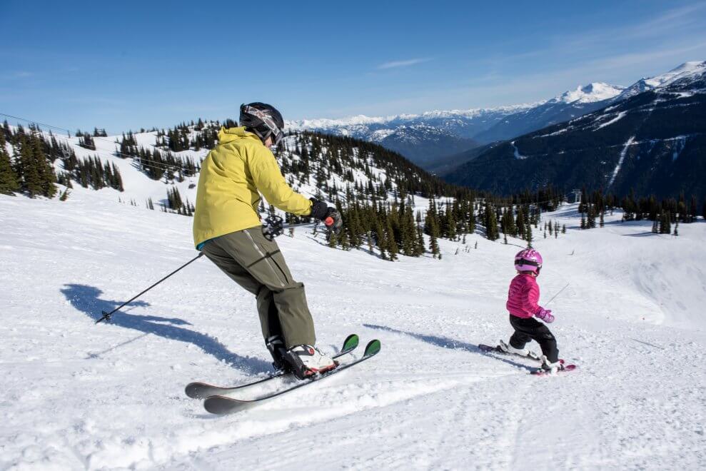 A dad and his young daughter ski down a slope at a ski resort.