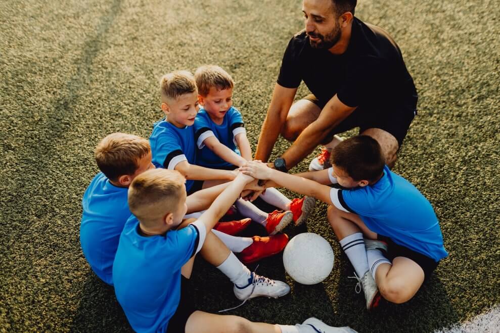 A boys' soccer team huddles and prepares for a cheer with their coach on the field.