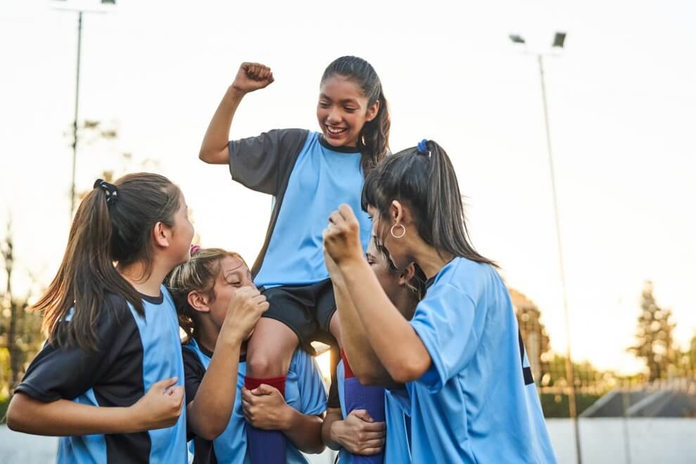 A girls' soccer team cheers on the field as they hoist one of the players onto two girls' shoulders.
