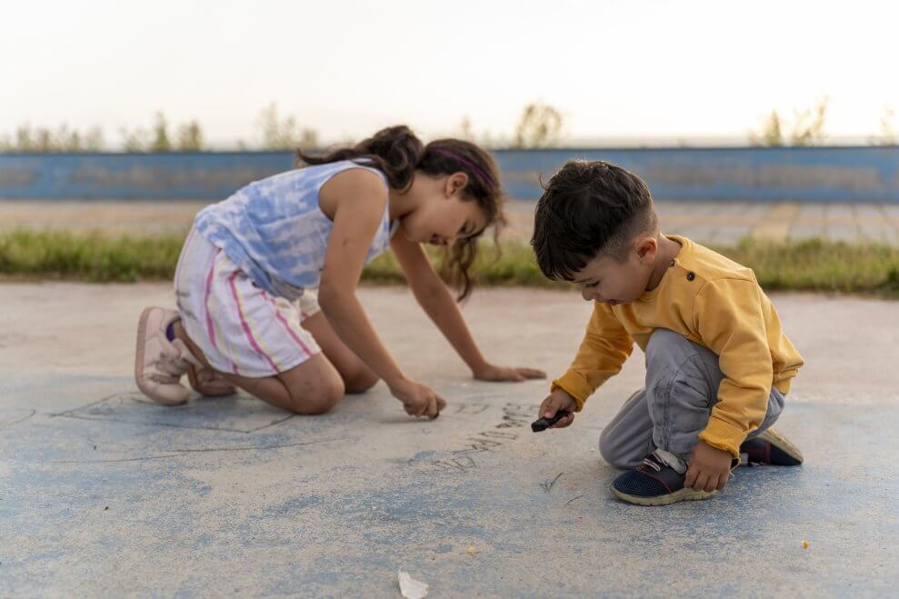 A toddler and his big sister draw with chalk on the sidewalk.
