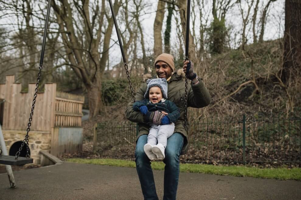 A father and his toddler swing at a playground together on a cold fall day. 