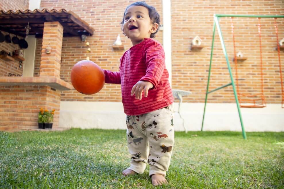 A smiling toddler stands barefoot in his backyard, clutching an orange ball.