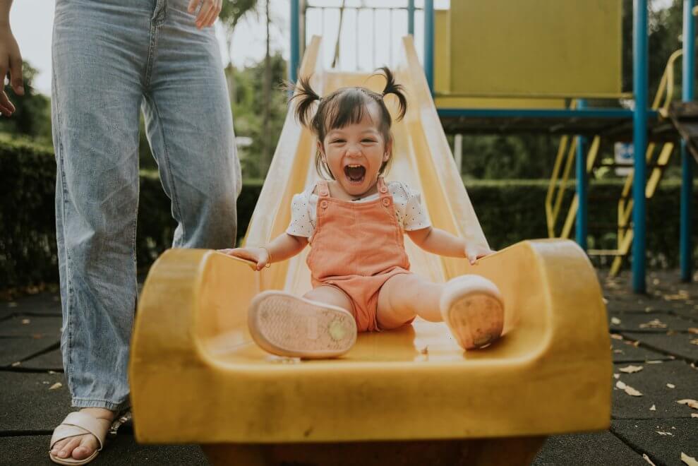A toddler shrieks with glee as she goes down a slide at a playground.