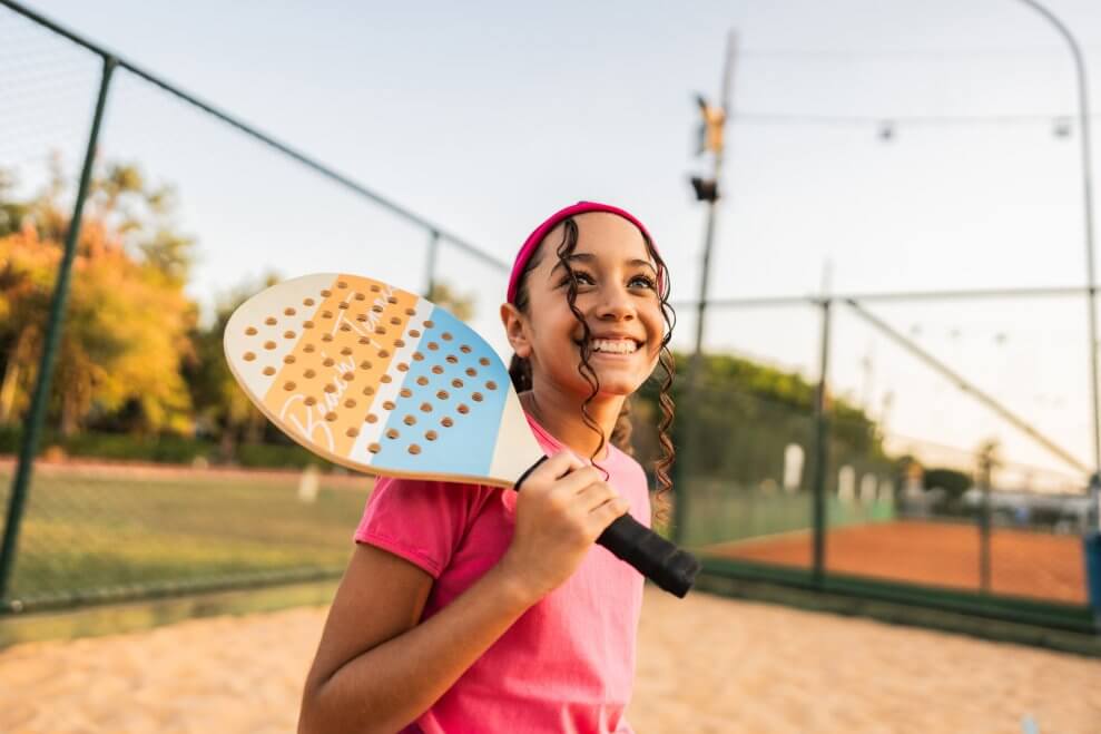 A girl stands on a beach court, holding a padel racquet.
