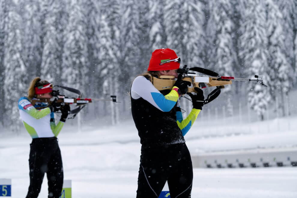 Two female biathlon athletes take aim with their rifles on a snowy course.