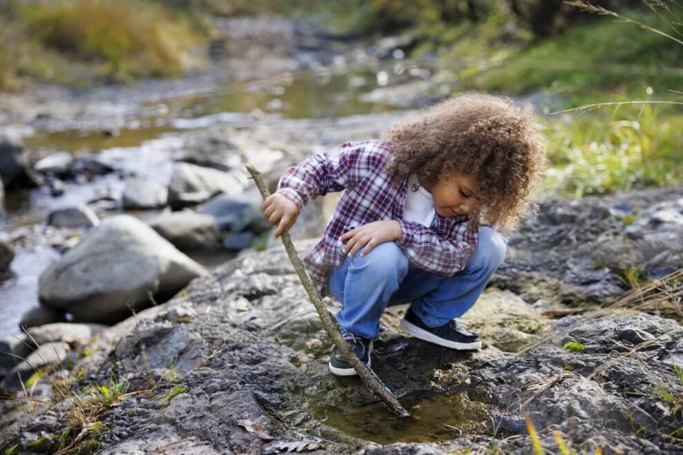 A child digs in the mud with a stick alongside a creek.
