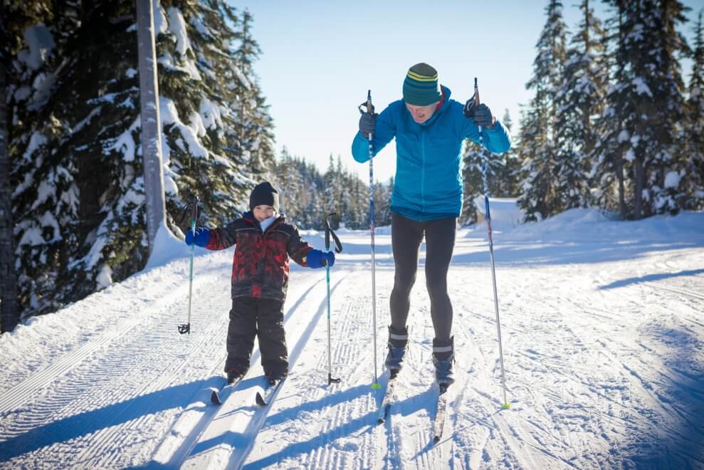 A dad and his young son cross-country ski together in the forest. The son has a smile on his face.