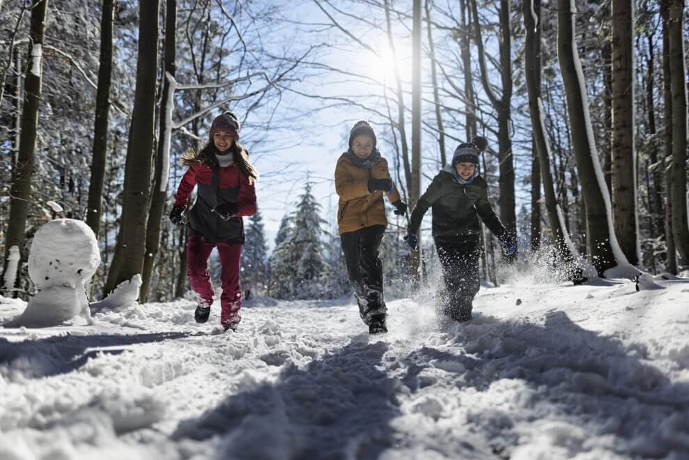 Three laughing kids run through a snowy forest on a sunny day.