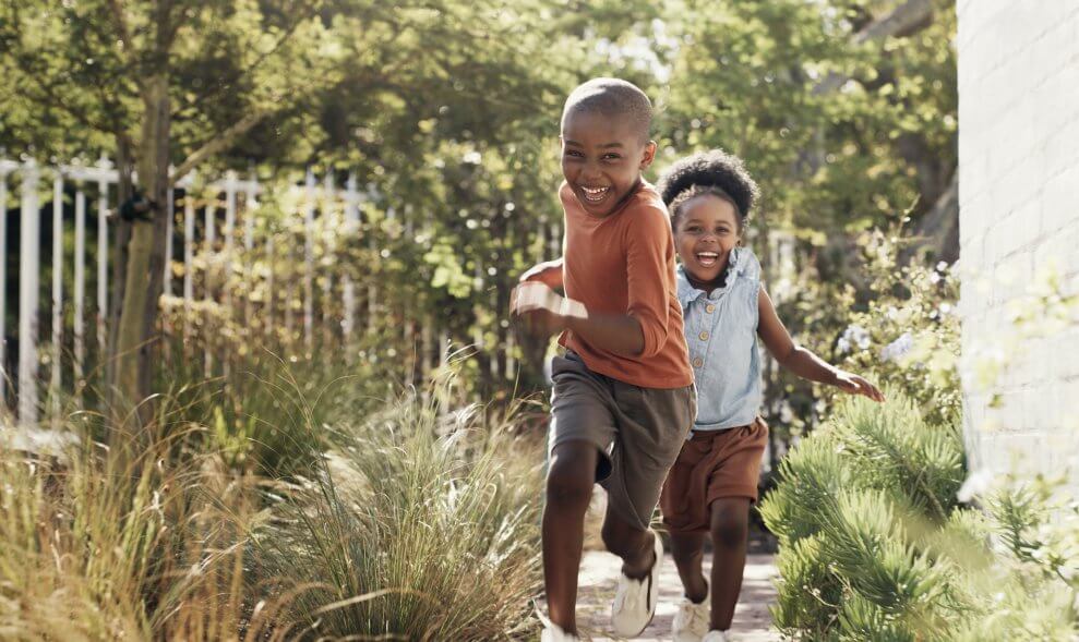 A brother and sister run around a garden together. They're both laughing.
