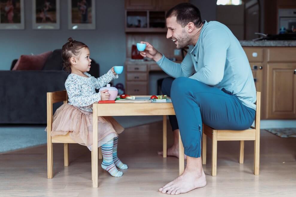 A dad and his toddler daughter sit at a child-sized table at home and do a toast with toy teacups.
