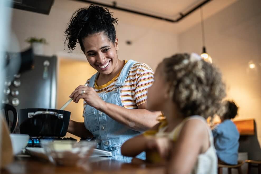 A mom smiles at her daughter as she stirs a pot on their stove in their kitchen.