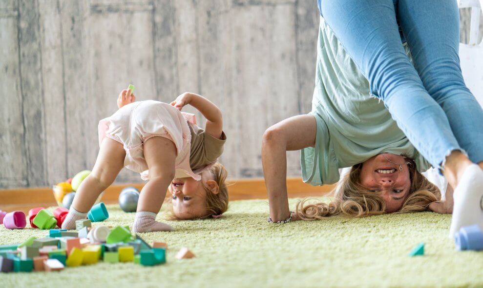A mother and her toddler play on the carpet at home together. With their feet resting on the ground, they both have their heads upside-down on the carpet.