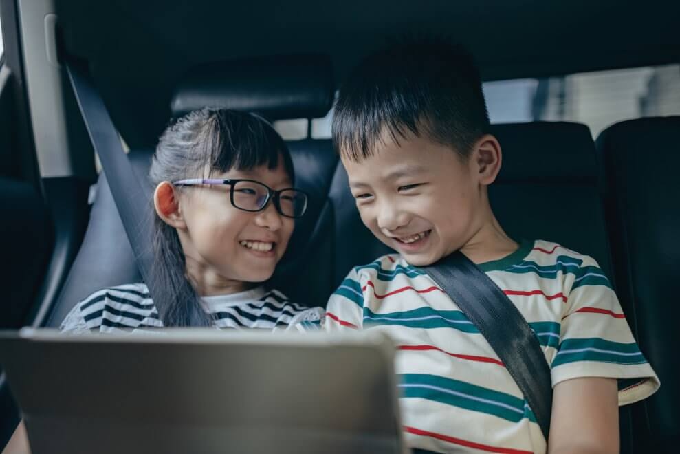 A brother and sister sit in the backseat of a car, using a tablet and laughing.
