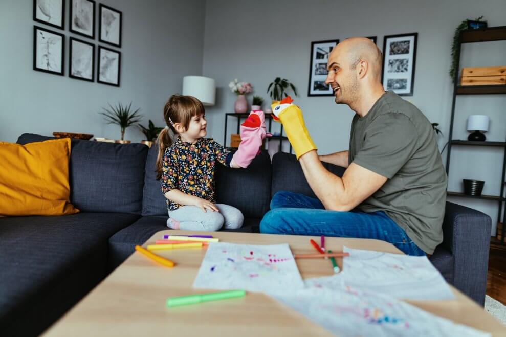 A dad and his young daughter play with puppets in the living room. 