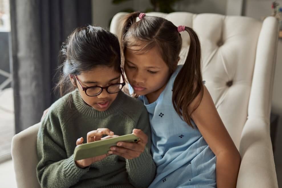 Two girls sit on an armchair at home and look at a smartphone together.