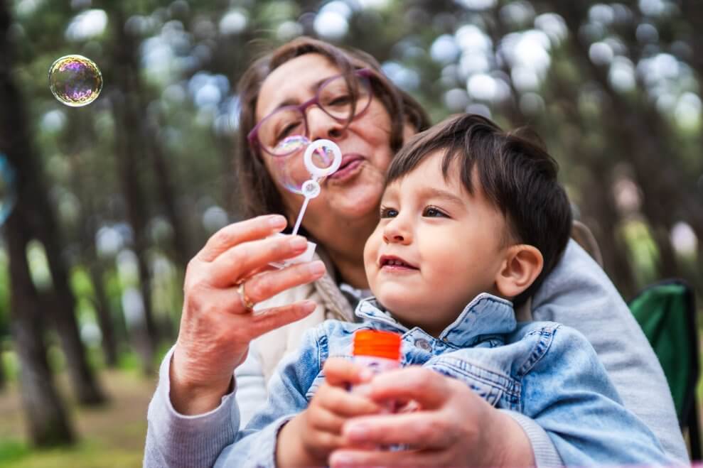 A grandma and her toddler grandson blow bubbles together in the yard.