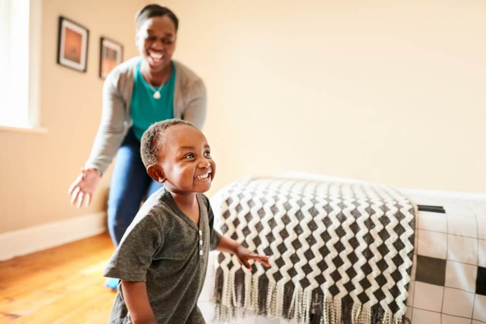 A mom chases her young son around the house. He has a big smile on his face.