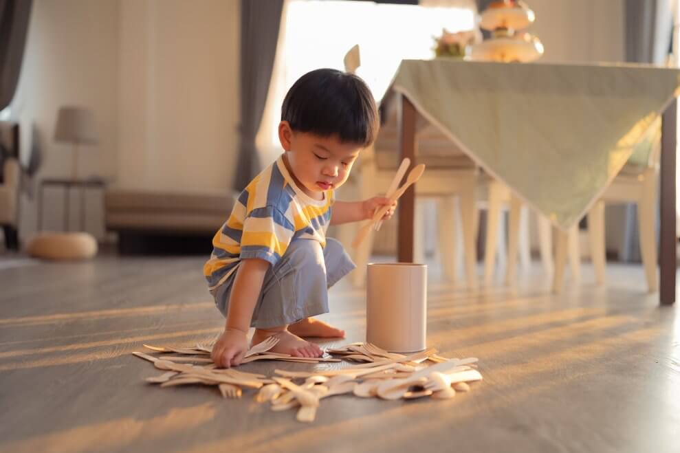 A toddler plays on the floor with a canister and wooden spoons and forks. This is one example of loose parts play.