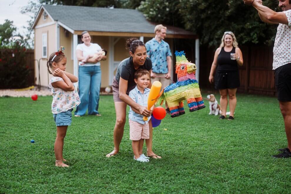 Families gather in a backyard as a young boy holds a bat and prepares to hit a pinata. His mom stands behind him, helping him aim. 