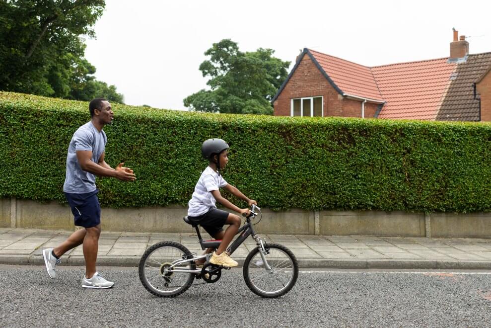 A dad teaches his son how to ride a bike on a quiet street. The son pedals ahead while his dad follows him, clapping.