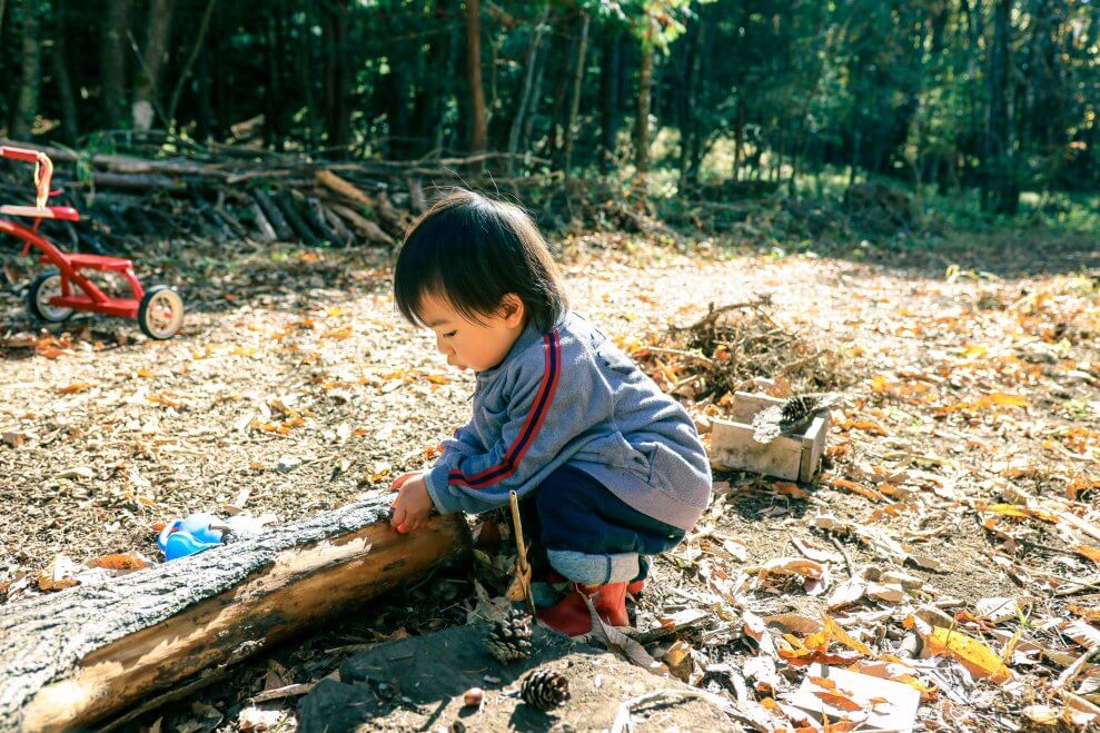 A young play crouches outside at a childcare centre and feels bark on a log.