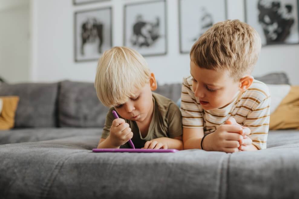 Two young brothers lie side by side on the couch, on their stomachs, and look at an tablet.