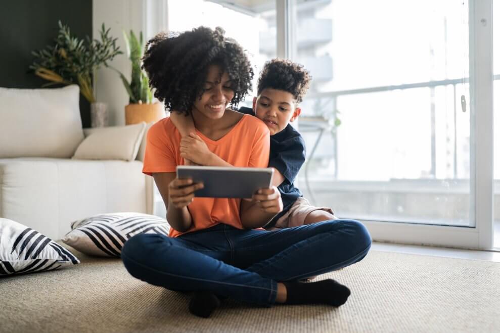 A teenage sister and her younger brother look at a tablet while they sit together on the living room floor at home.