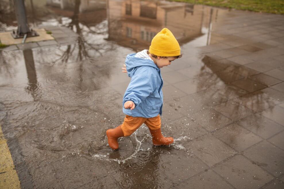 A toddler wearing a toque, rain jacket, and rubber boots splashes through a puddle.