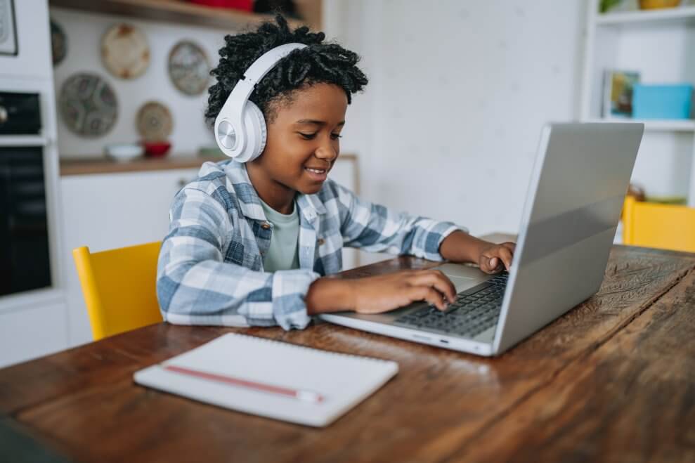A boy sits at the kitchen table at home, wearing headphones and doing homework on a laptop. A notebook and pencil sit on the table beside him.