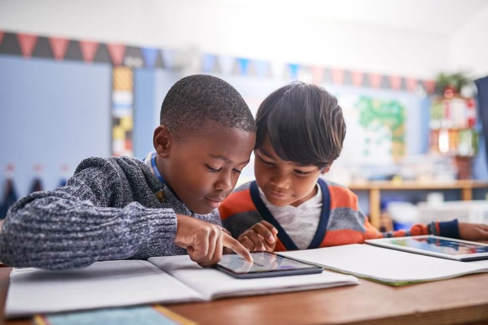 Two boys sit at a desk in a classroom and use a tablet together.