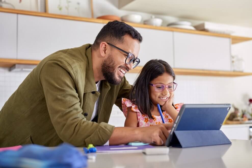 A daughter sits at the kitchen counter in her house, using  tablet to do her homework. Her dad stands next to her, helping her with it.