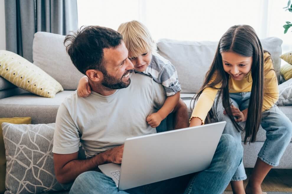 A dad and his young and son and daughter sit in their living room and look at a laptop that sits on the dad's lap.