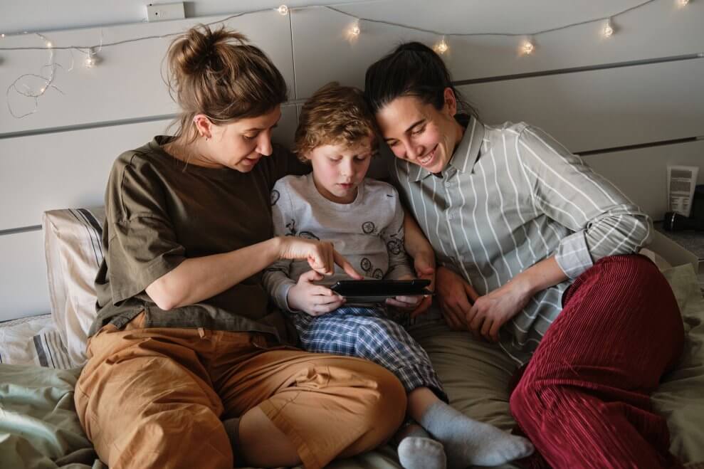 Two moms and their elementary-age son sit together at home and look at a tablet screen together.