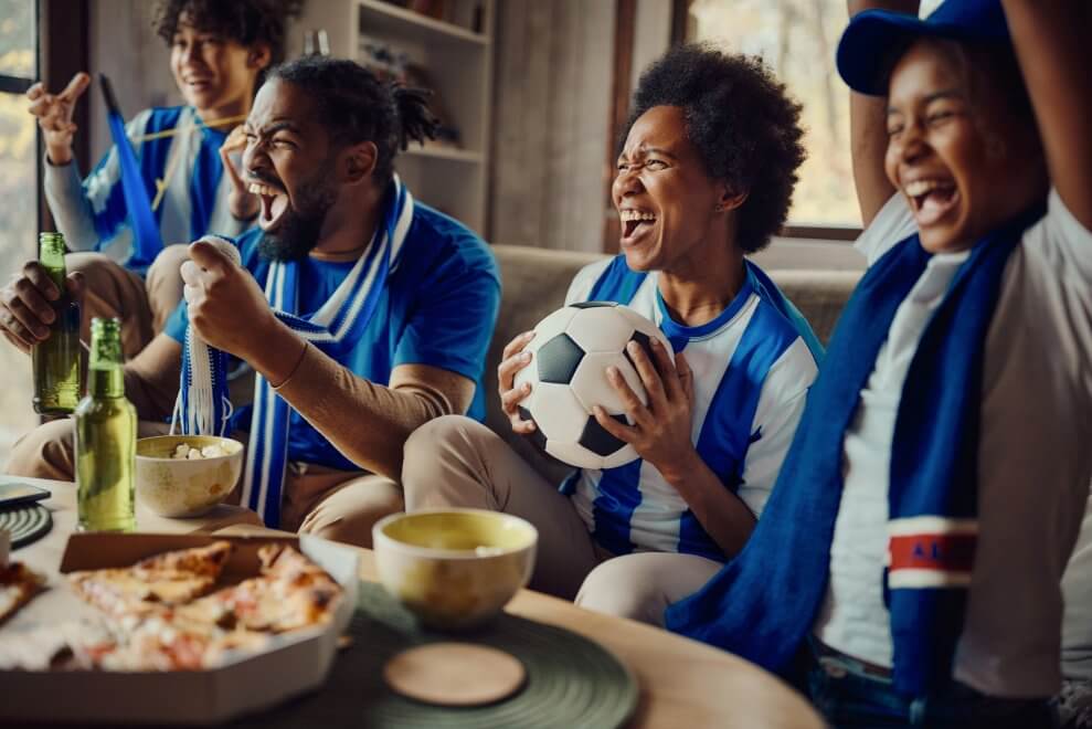 A mom, dad, son, and daughter watch a soccer game on TV at home. They sit on the couch together, eating pizza and cheering.