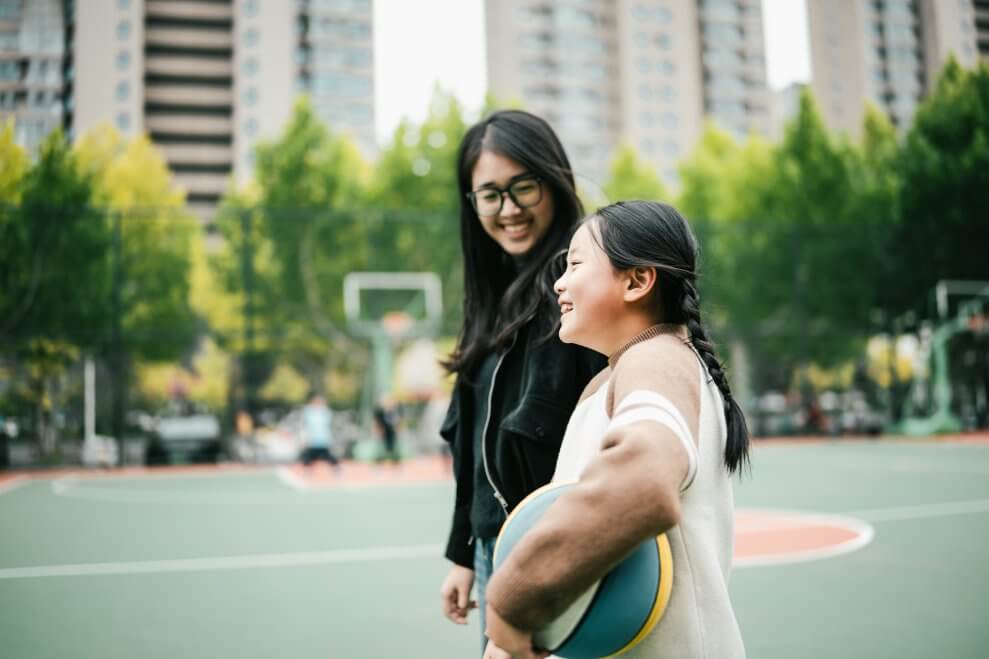 A mom and her young daughter play basketball on an outdoor court in ab urban neighbourhood.