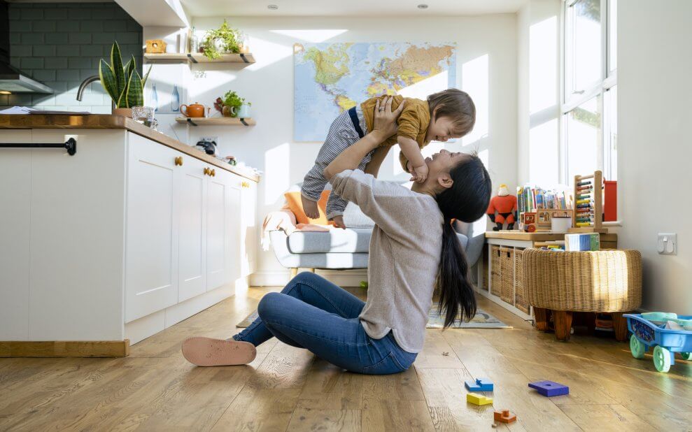 A smiling mom sits on the floor in her house, holding her toddler up in the air. The toddler smiles back at her