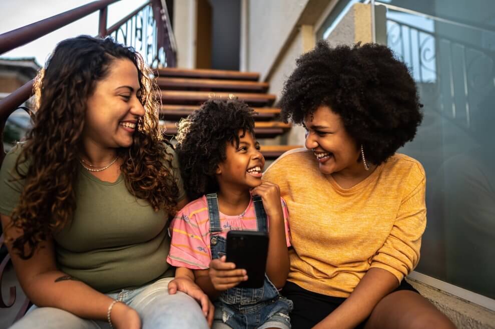 Two moms sit with their daughter on a set of stairs outdoors. The daughter holds a phone in her hand, and all three of them are smiling at each other.