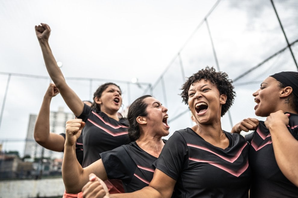 Five members of a women's soccer team shout and celebrate on an outdoor pitch.