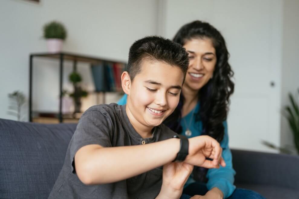 A preteen boy looks at his smartwatch while his mom sits next to him on the couch at home.