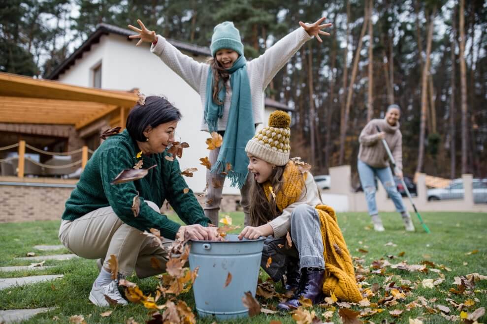 Two girls and their grandmother make leaf soup in a large bucket. Behind them, their mother rakes leaves.