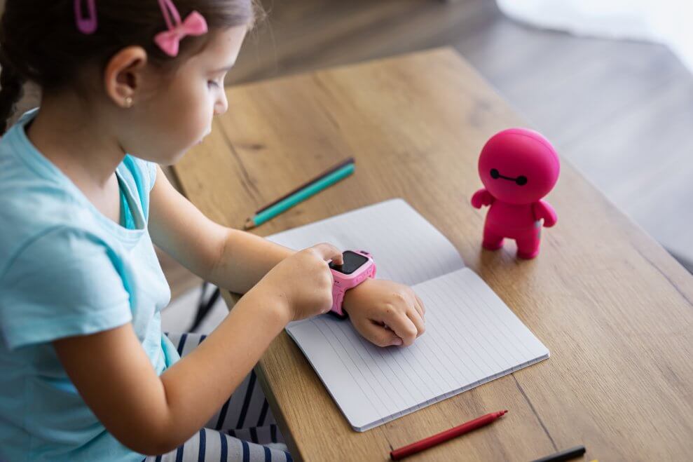 A young girl wears a smartwatch and taps on the screen while she's sitting at a desk with a notebook in front of her.