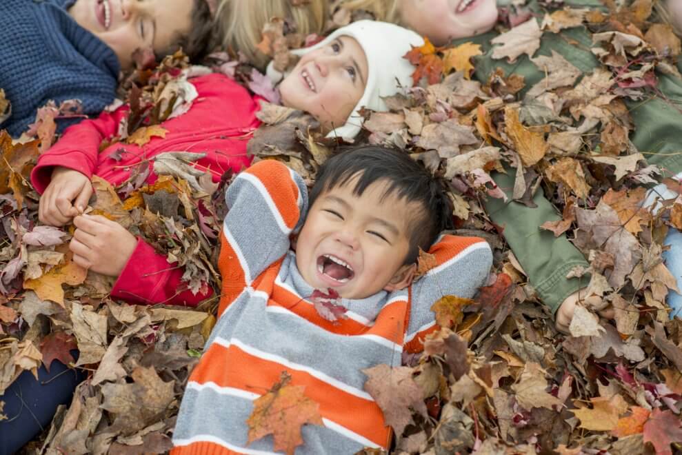 A group of laughing young kids lie in a pile of orange and brown leaves.