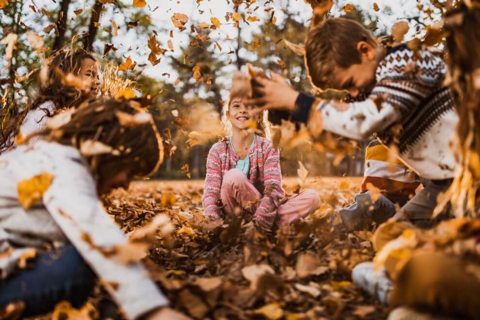 A group of elementary-age kids play outside in leaf pile. They're toss leaves into the air and laughing as they fall down.