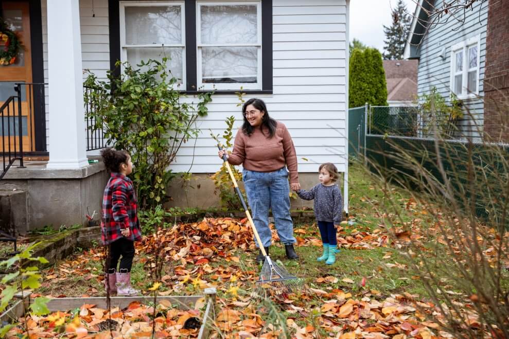 A mom and her two young daughters stand in their leaf-covered front yard. The mom holds a rake and the hand of her toddler daughter.