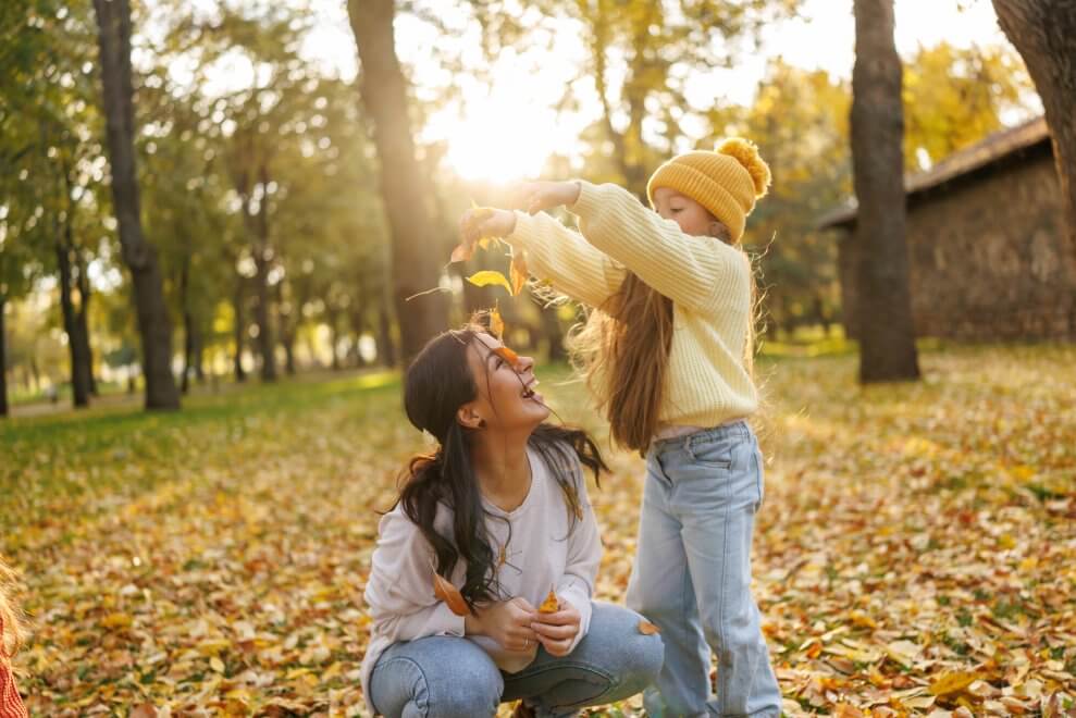 An elementary-age girl tosses leaves onto her mom's head. The two are playing outside on a sunny fall day.