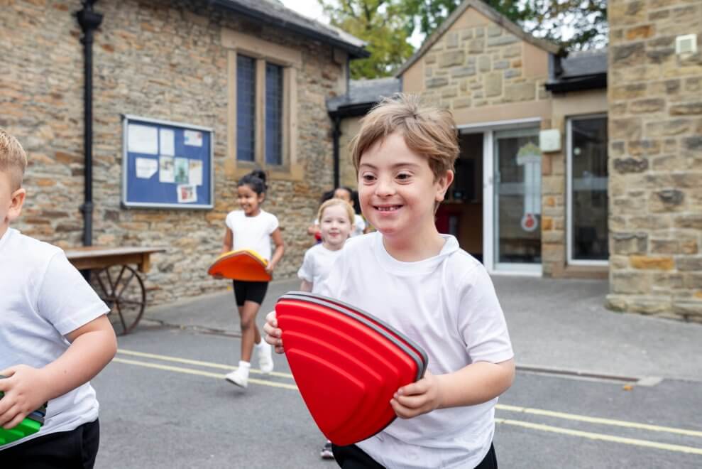 A boy with Down syndrome carries a red sports cone outside for phys ed class. His fellow classmates follow him.